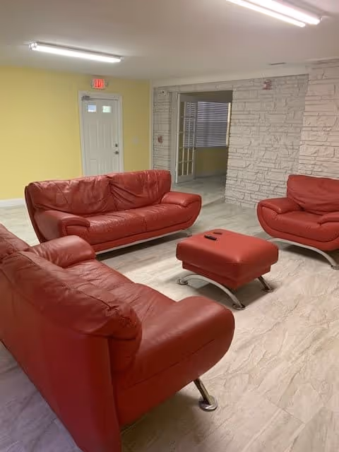 A living room area with three red leather sofas and a matching red ottoman on a light-colored tiled floor. The walls are painted yellow and white with a stone texture on one side. There is a white door with a window and an exit sign above it, and fluorescent ceiling lights illuminate the room.