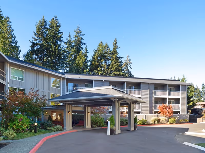 Front entrance and porte-cochere of a three-story assisted living building with balconies, landscaping, and tall trees under a clear blue sky.