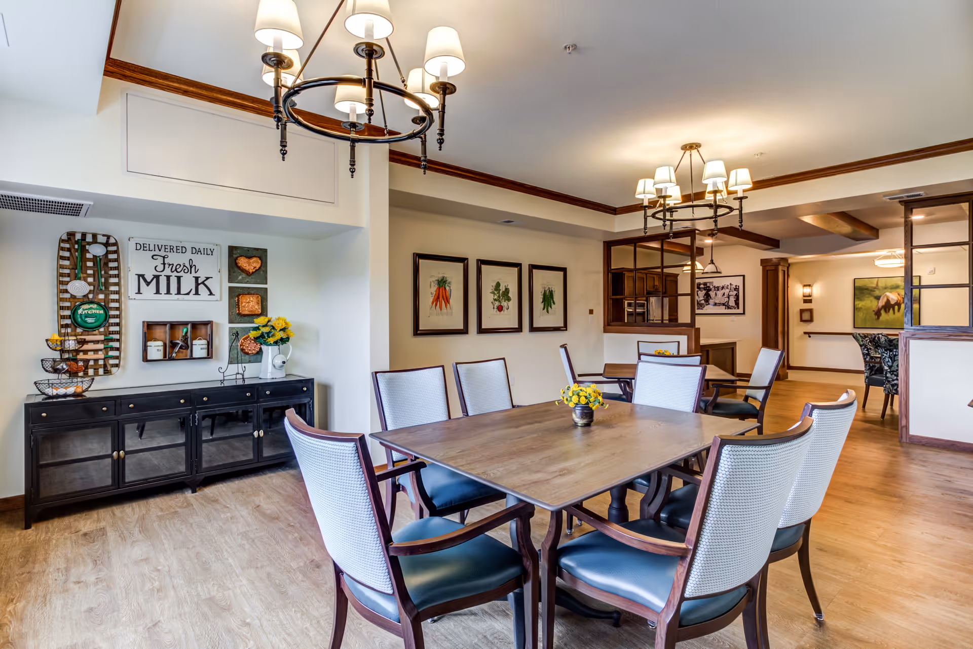 A bright and inviting dining room in a senior living facility with a wooden table surrounded by six cushioned chairs. The room features wooden flooring, decorative wall art including framed vegetable prints and a sign that reads 'Delivered Daily Fresh Milk'. There are two chandeliers with lampshades hanging from the ceiling, and a black sideboard with decorative items such as a vase with yellow flowers and kitchen-themed decor on the wall.