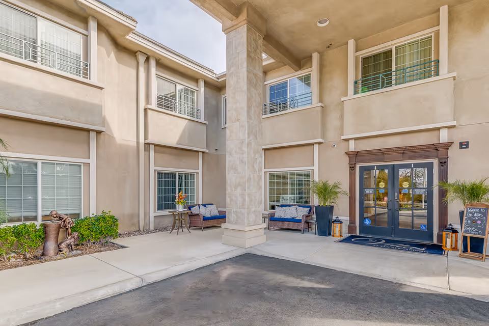 Covered front entrance of a senior living facility with double glass doors, seating areas, potted plants, and a large central pillar.