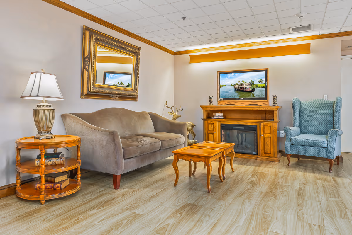 A cozy living room area featuring a gray upholstered sofa, a wooden side table with a decorative lamp and books, a wooden coffee table, a wooden electric fireplace with a TV mounted above it displaying a boat on water, and a blue upholstered armchair. The room has light wood flooring, a large framed mirror on the wall, and neutral-colored walls with crown molding.