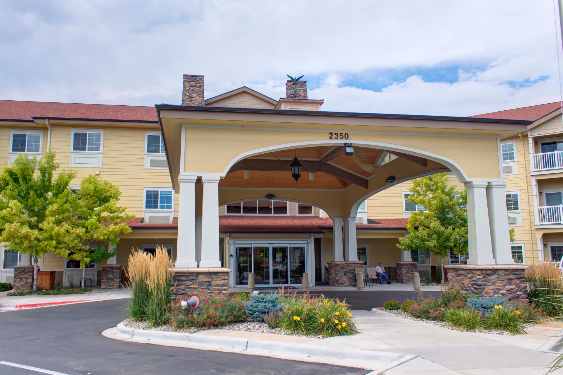 Front entrance of Rigden Farm Senior Living facility with a covered drop-off area supported by columns. The building is three stories tall with yellow siding and red roofing. There are landscaped plants and trees around the entrance, and a person is sitting on a bench near the door.