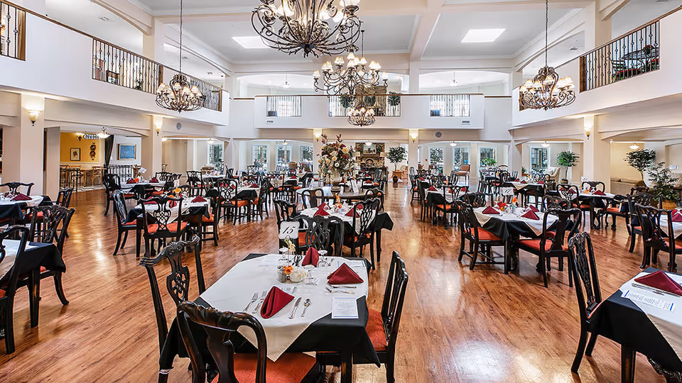 Spacious senior living dining room with multiple tables set with white and black tablecloths, red napkins, and silverware. The room features wooden floors, elegant chandeliers hanging from a high ceiling, and a second-floor balcony overlooking the area. There are plants and decorative elements around the room, creating a warm and inviting atmosphere.