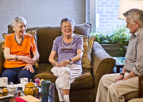 Three elderly people sitting and smiling in a cozy living room. Two women are seated on a brown couch with decorative pillows, and a man is sitting on a chair nearby. A coffee table with cups, a teapot, and other items is in front of them. A window with plants outside is visible in the background.