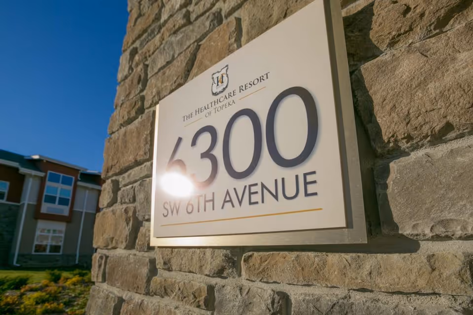Close-up view of a stone wall with a mounted sign displaying the address '6300 SW 6th Avenue' and the name 'The Healthcare Resort of Topeka' under a clear blue sky.