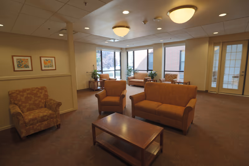 A spacious living room area in a senior living facility with multiple upholstered armchairs and sofas arranged around wooden coffee tables. Large windows allow natural light to fill the room, and there are framed artworks on the walls and potted plants near the windows. The ceiling has recessed lighting and two round ceiling lights.