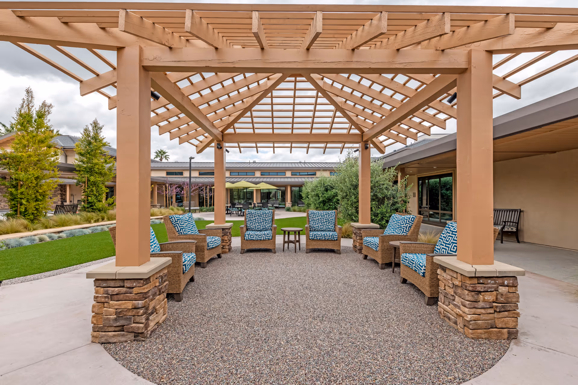 Outdoor seating area under a wooden pergola with six cushioned wicker chairs arranged in a semi-circle around two small tables, surrounded by greenery and buildings in the background under a cloudy sky.