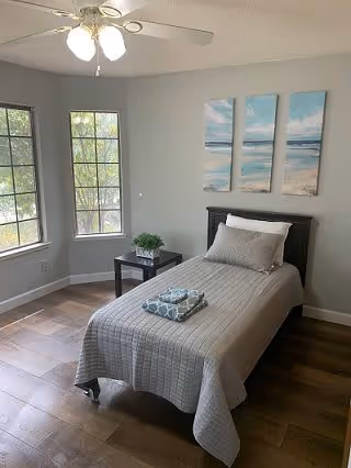 A simple, clean bedroom with a single bed covered in a gray quilt and a folded towel on top. There is a small black bedside table with a green plant, two large windows letting in natural light, a ceiling fan with lights, and a three-panel beach-themed artwork on the wall above the bed.