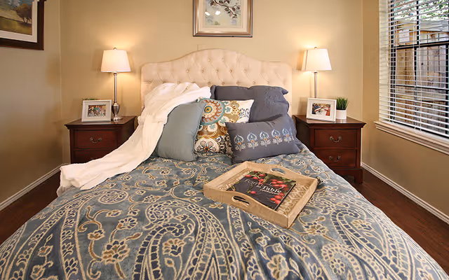 A cozy bedroom with a bed featuring a tufted cream-colored headboard, blue and patterned pillows, and a blue paisley-patterned bedspread. There is a wooden tray with a book on the bed. Two matching wooden nightstands flank the bed, each with a lamp, a framed photo, and a small plant. A window with blinds is visible on the right side, and a framed picture hangs above the headboard.