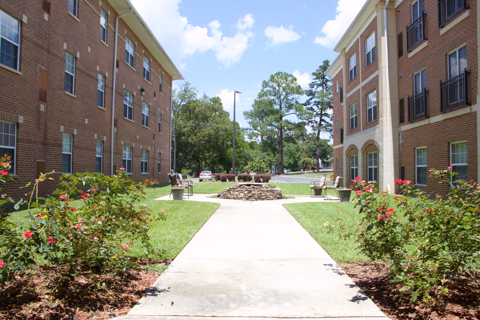 Outdoor courtyard area between two brick buildings with a concrete walkway leading to a circular stone fire pit surrounded by chairs. There are green lawns, flower bushes with pink and red flowers, and trees in the background under a partly cloudy blue sky.