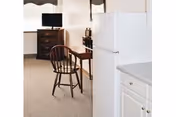 Interior view of a room showing a white refrigerator and kitchen cabinets in the foreground, with a wooden chair, a small wooden desk, a dresser with a television on top, and a window with curtains in the background.