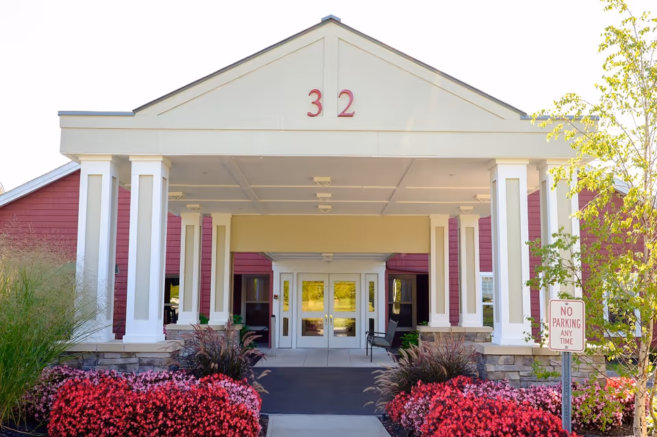 Covered entrance and portico of a red-sided senior living building with white columns, the number 32 above the doorway, and flowering landscaping.