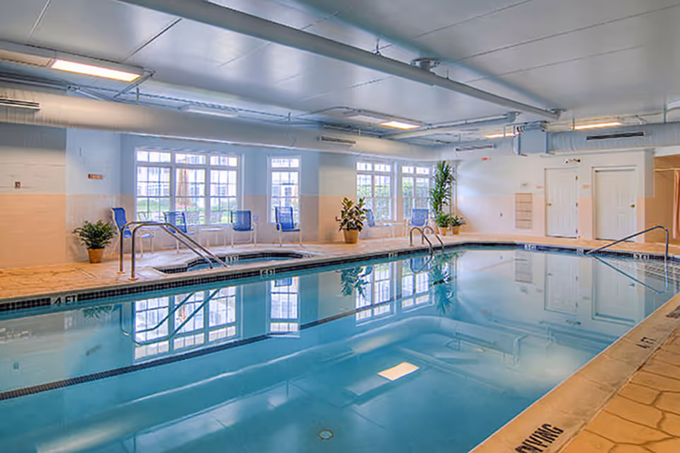 Indoor swimming pool and adjacent spa in a bright recreation room with chairs, potted plants, and large windows.