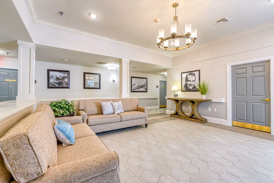 A bright and spacious senior living facility common area with two beige sofas adorned with decorative pillows, a wooden console table with a lamp and a potted plant, framed black and white photographs on the walls, a modern chandelier hanging from the ceiling, and gray doors with gold handles.
