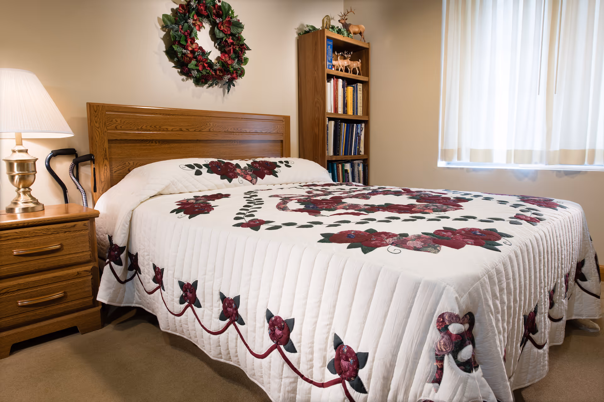 A cozy bedroom with a wooden bed frame and a bed covered with a white quilt featuring red and green floral patterns. A wooden nightstand with a brass lamp and a cane is beside the bed. A wooden bookshelf filled with books and decorative items stands against the wall. A festive wreath hangs above the bed, and a window with white curtains allows natural light into the room.