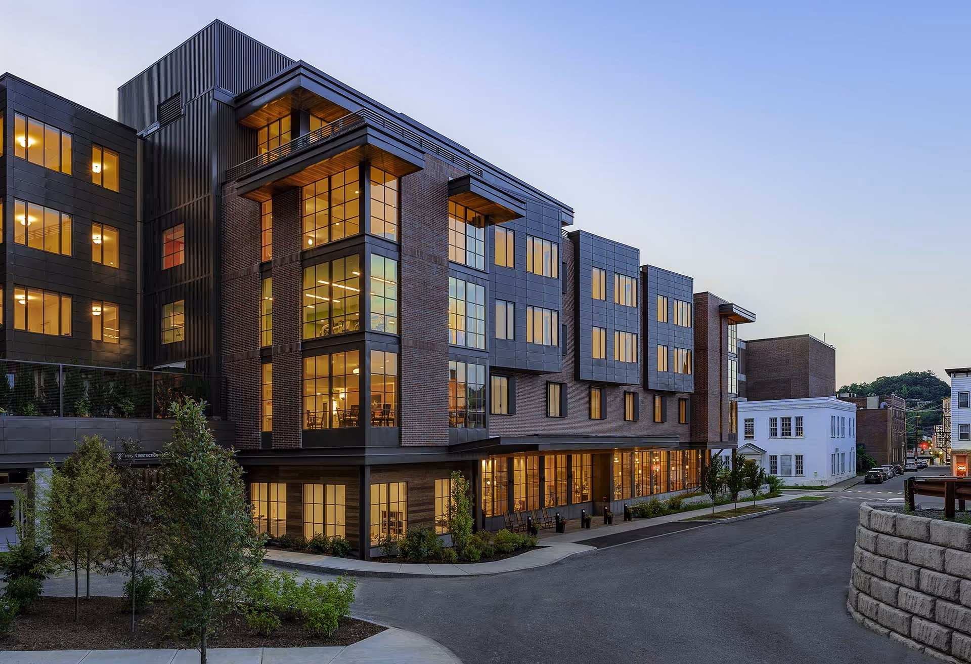 Modern multi-story building with large illuminated windows and a brick-and-metal facade on a quiet street at dusk.