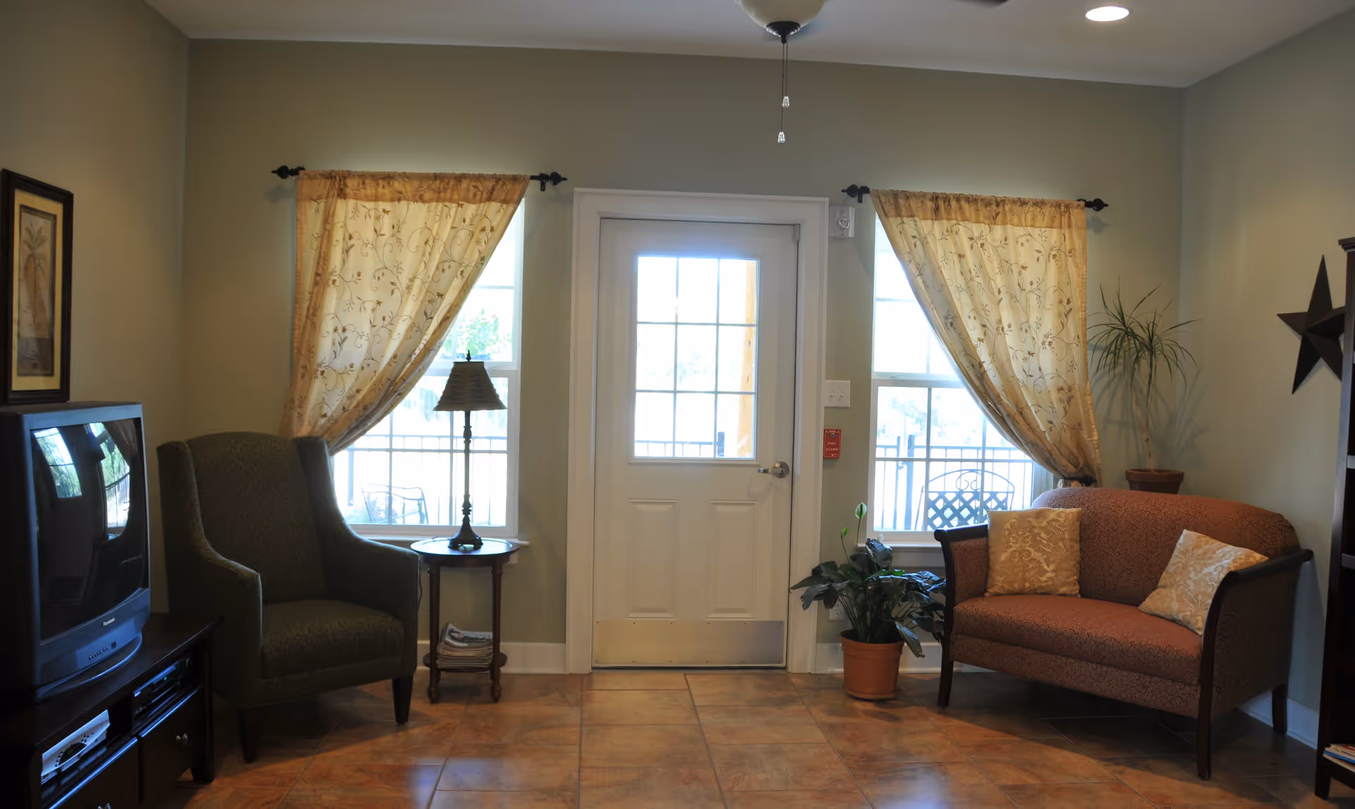 Cozy living room with an armchair, loveseat, side table and lamp, potted plants, and a central door flanked by curtained windows.