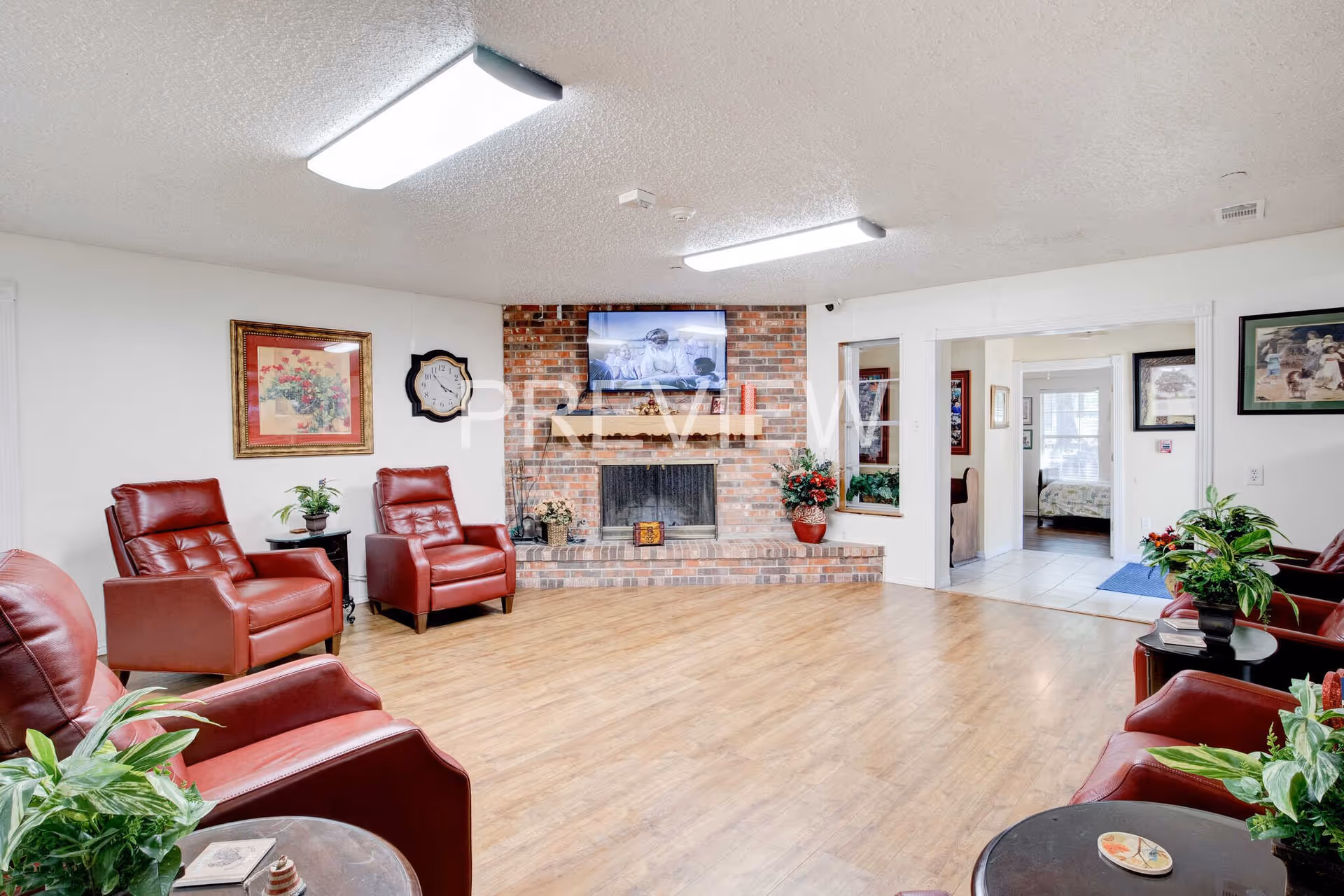 A cozy living room area with six red leather armchairs arranged around the perimeter. The room features a brick fireplace with a flat-screen TV mounted above it. There are several potted plants on small tables and on the fireplace hearth. The walls are white with framed artwork and a clock. The floor is light wood, and there is an open doorway leading to another room with a bed visible.