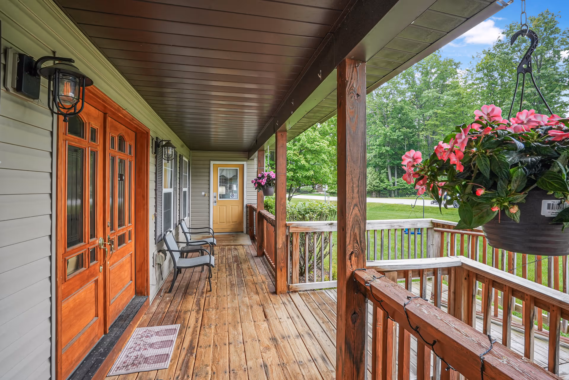 A wooden porch with a wooden floor and railing, two black metal chairs with gray cushions, a hanging flower pot with pink flowers, and two doors: a double wooden door on the left and a single yellow door at the end. The porch overlooks a green lawn and trees under a partly cloudy sky.