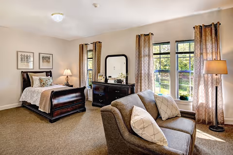 A well-lit bedroom in a senior living facility featuring a wooden bed with white and beige bedding, a matching dresser with a mirror, two framed artworks on the wall, a cozy gray loveseat with patterned pillows, and three large windows with beige curtains letting in natural light. A floor lamp stands near the windows.
