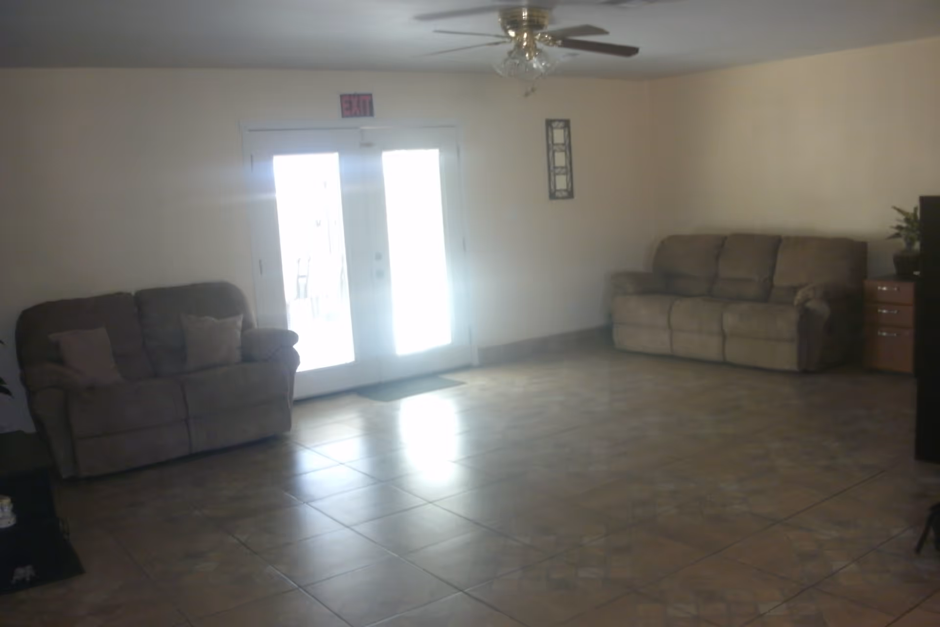 A living room with two beige sofas placed against opposite walls, a ceiling fan with lights, tiled floor, and double glass doors with an exit sign above them letting in bright light.