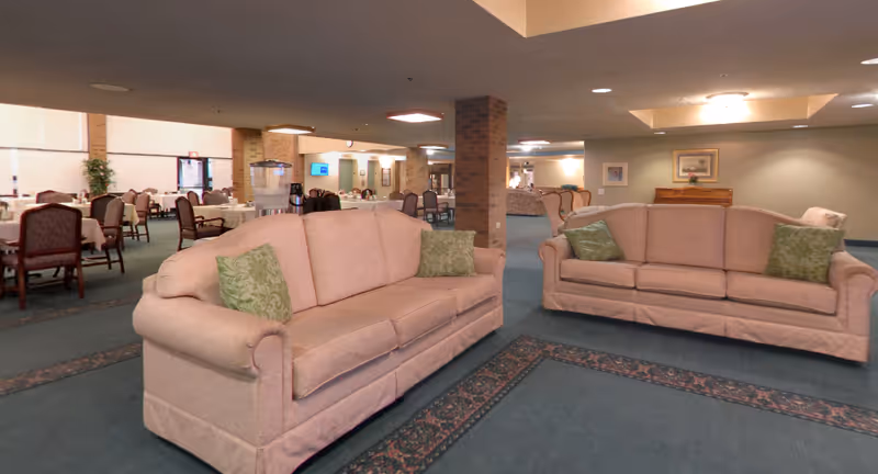 A spacious senior living common area with two beige sofas adorned with green patterned pillows. In the background, there are multiple dining tables with chairs, a water dispenser, and a few framed pictures on the walls. The room has a carpeted floor with patterned borders and brick pillars supporting the ceiling.