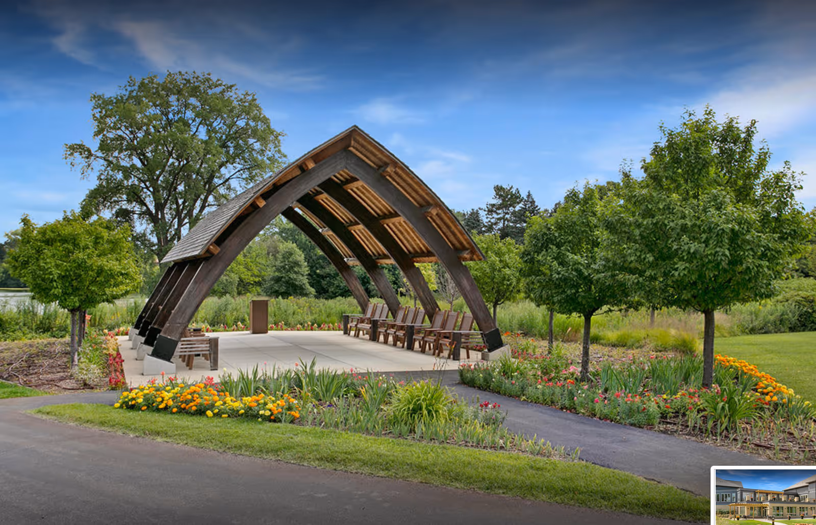 Outdoor pavilion with a curved wooden roof structure and several wooden chairs underneath, surrounded by landscaped gardens with colorful flowers and trees, set against a blue sky with some clouds.
