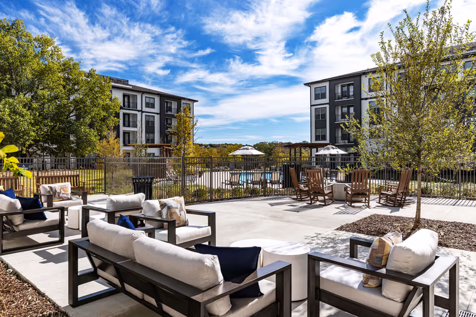 Outdoor patio area with modern cushioned seating and wooden chairs arranged around small tables, surrounded by trees and landscaping. In the background, there are multi-story residential buildings under a partly cloudy blue sky.