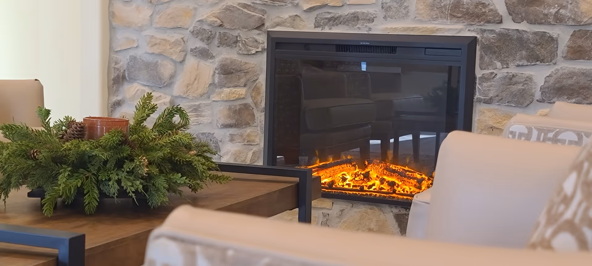 Cozy living room area with a stone fireplace featuring a glowing electric fire, surrounded by beige upholstered chairs and a wooden coffee table decorated with a green pine and pinecone centerpiece.