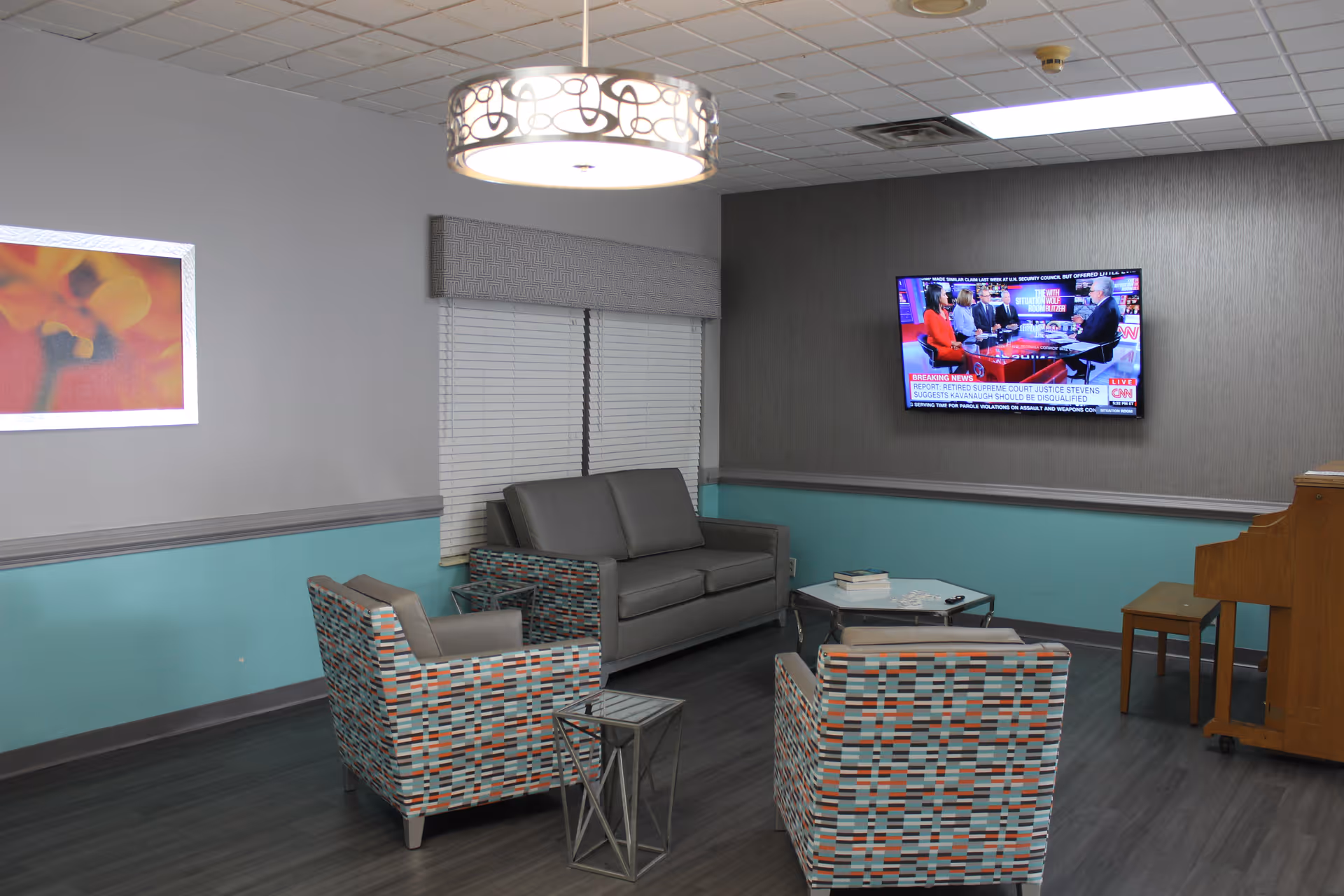A cozy living room area with a gray couch, two patterned armchairs, a small side table, and a glass coffee table. A flat-screen TV is mounted on the wall showing a news program. The walls are painted gray and teal, with a window covered by blinds and a valance. A piano and piano bench are visible on the right side of the room. A decorative ceiling light fixture hangs above.