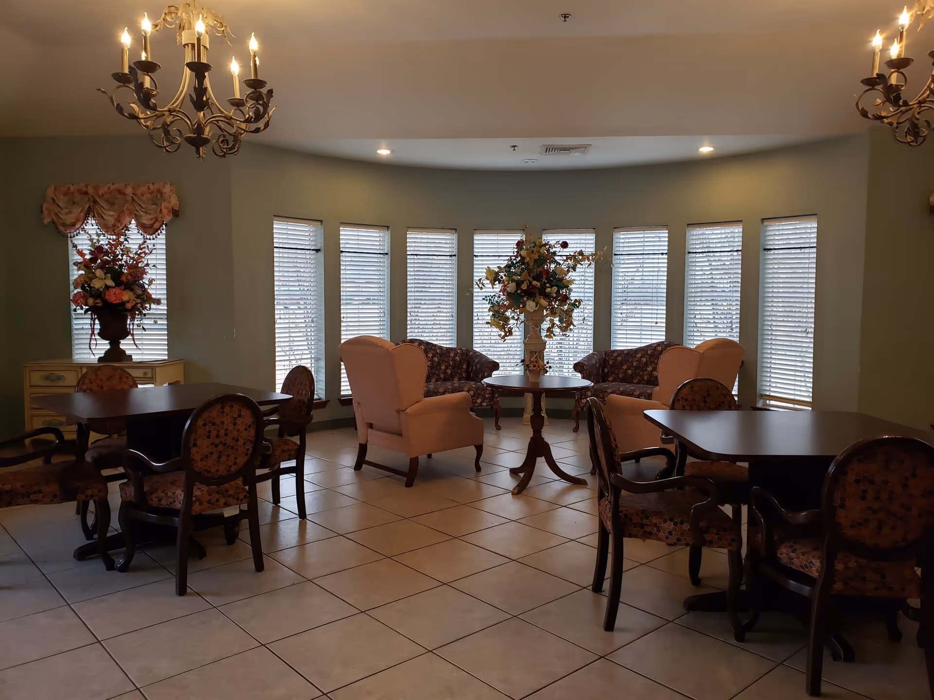 A cozy sitting area with multiple tables and chairs arranged on a tiled floor. The room features two chandeliers hanging from the ceiling, large windows with blinds, and floral arrangements on tables and a side cabinet. The walls are painted a soft green color.
