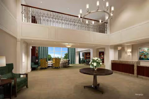 Spacious and elegant senior living facility lobby with a round table holding a flower arrangement in the center, green upholstered chairs to the left, a reception desk to the right, and a mezzanine with decorative railing above. Large windows in the background let in natural light and offer a view of greenery outside.