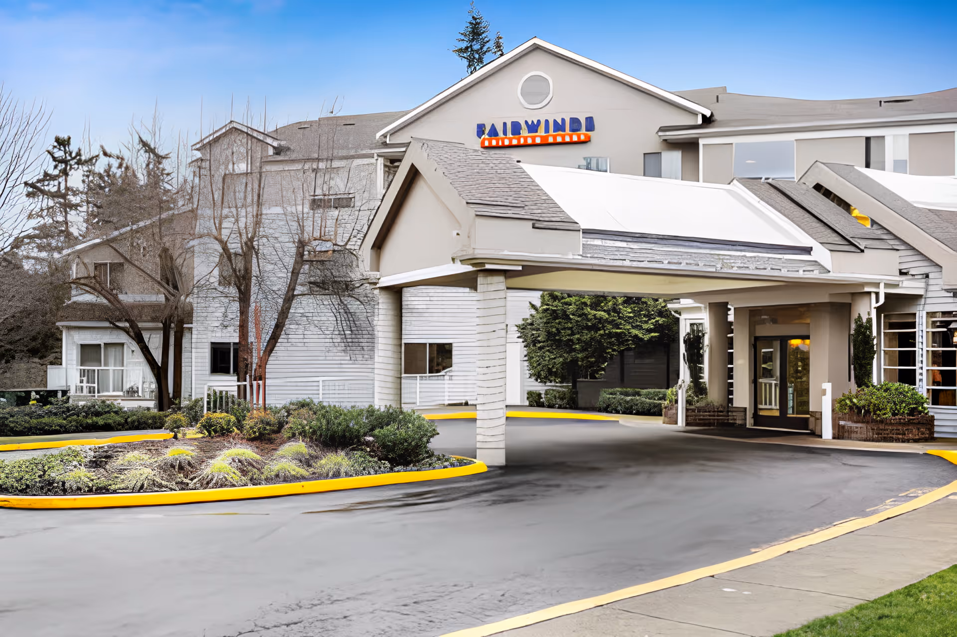 Exterior view of Fairwinds - Brighton Court senior living facility showing the main entrance with a covered driveway, landscaped garden beds, and a clear blue sky.