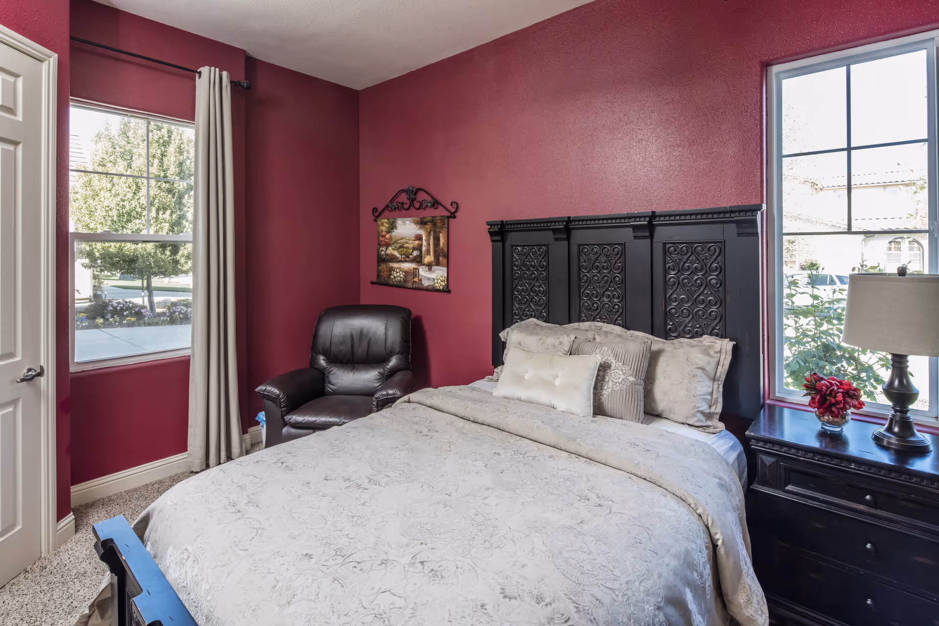 A bedroom with dark red walls featuring a large bed with a decorative black headboard and beige bedding. There is a black leather armchair in the corner, a nightstand with a lamp and a small vase of red flowers, and two windows letting in natural light.