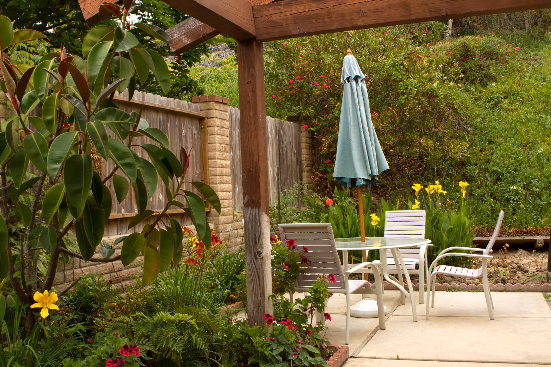 Outdoor patio area with a wooden pergola, a glass-top table with four white chairs, and a closed blue umbrella. The patio is surrounded by a garden with various green plants and colorful flowers, and a wooden fence with brick pillars in the background.