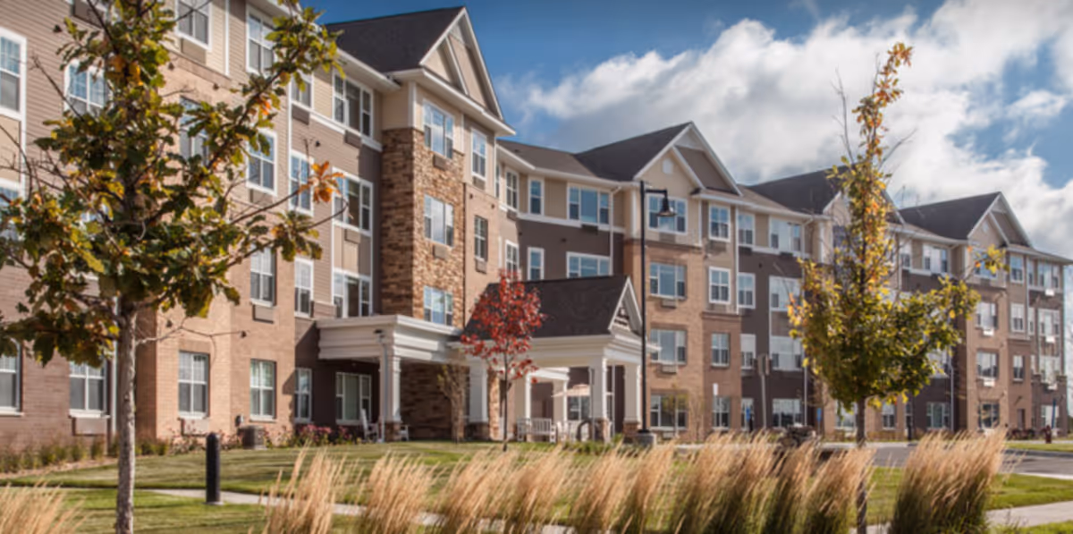 Exterior view of a multi-story senior living facility building with a covered entrance, surrounded by trees and landscaping under a partly cloudy sky.