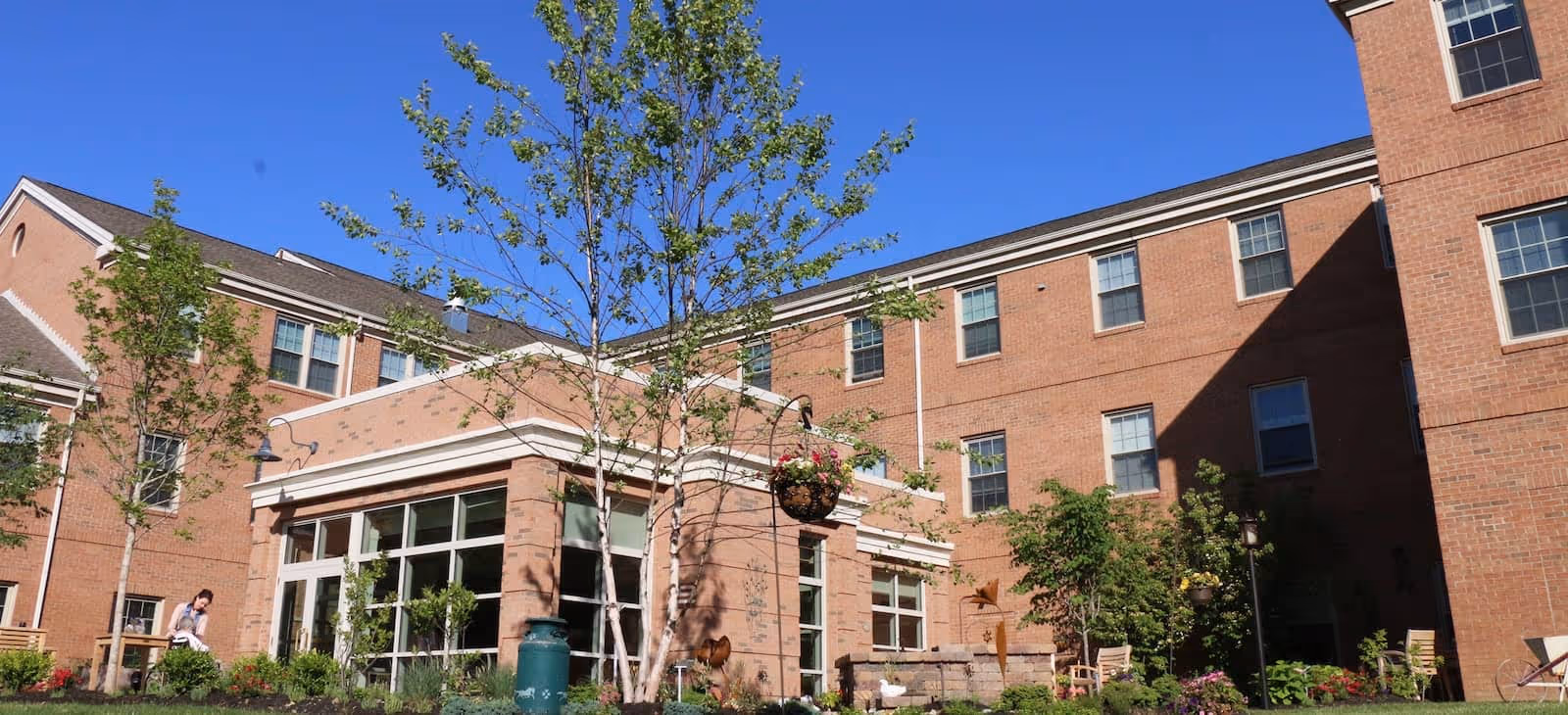 Outdoor courtyard area of a senior living facility with brick buildings surrounding it, trees, hanging flower baskets, and garden landscaping under a clear blue sky.