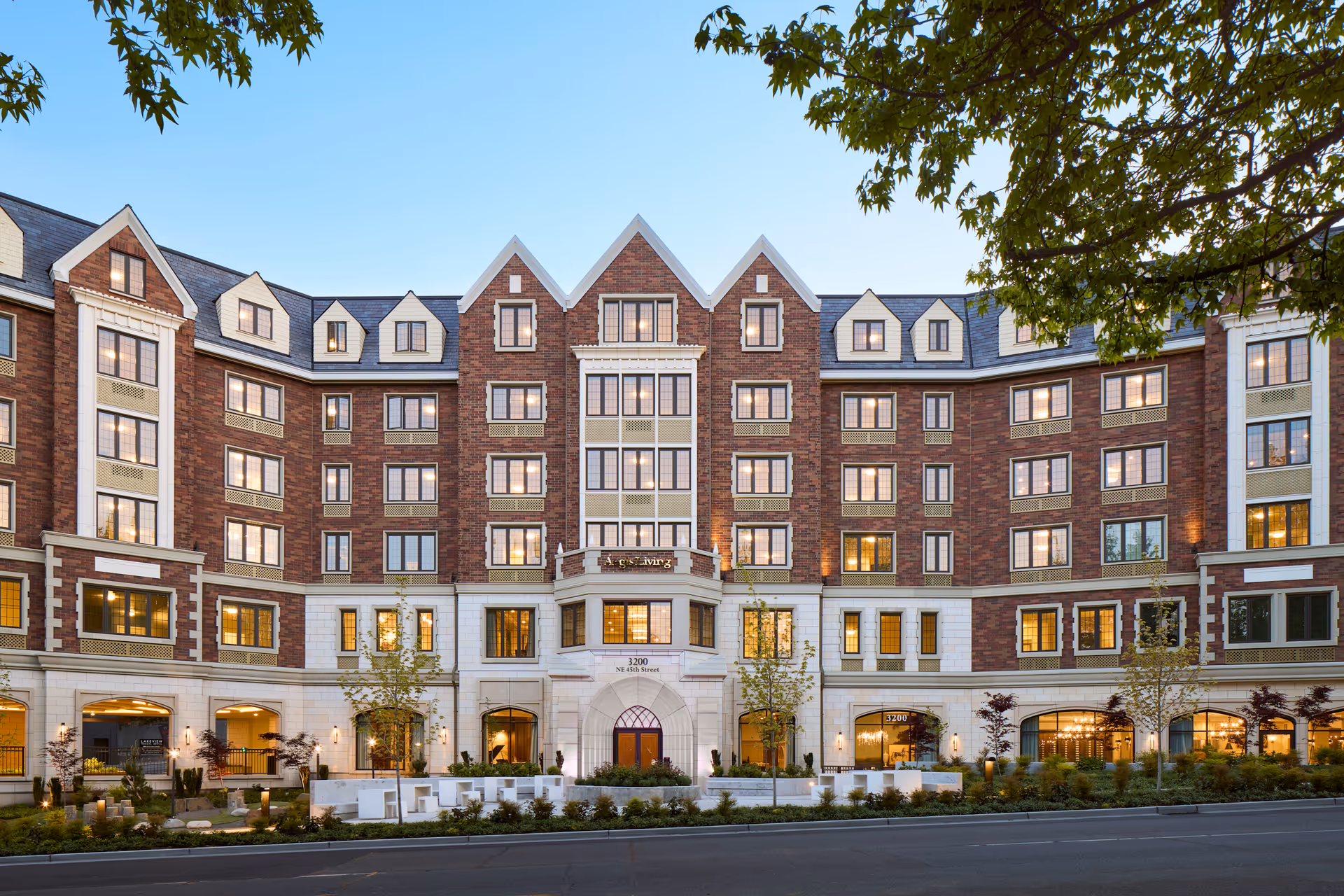 Front exterior view of a large multi-story senior living facility building with brick and white stone facade, multiple windows lit from inside, and landscaped greenery in front under a clear blue sky.