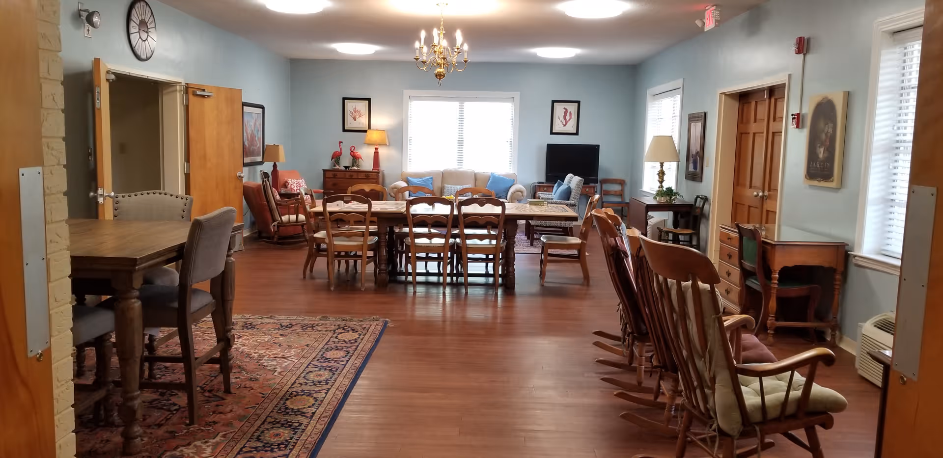 Spacious common room with a long dining table surrounded by chairs, rocking chairs along one side, and sofas and lamps near the windows.