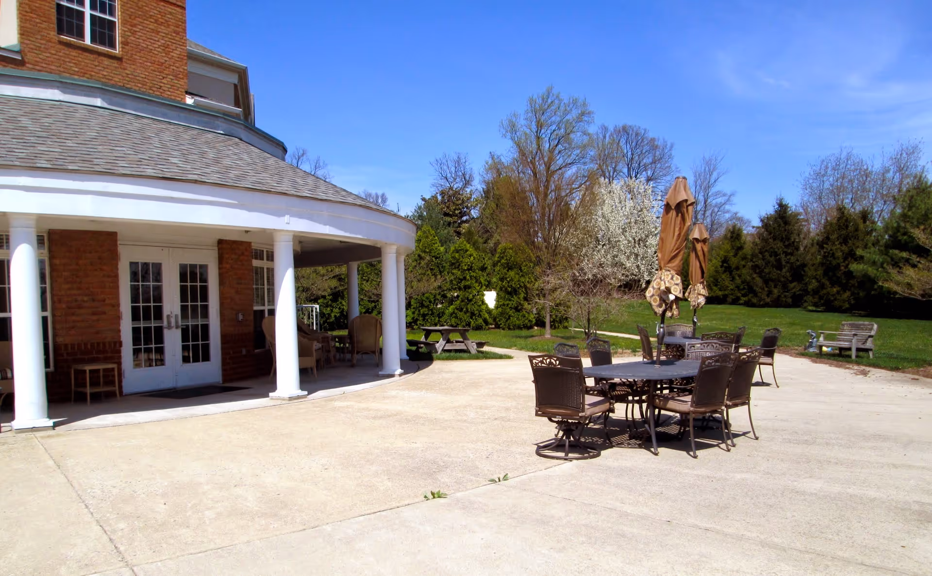 Outdoor patio with metal tables, chairs and closed umbrellas beside a brick building with white columns and French doors.