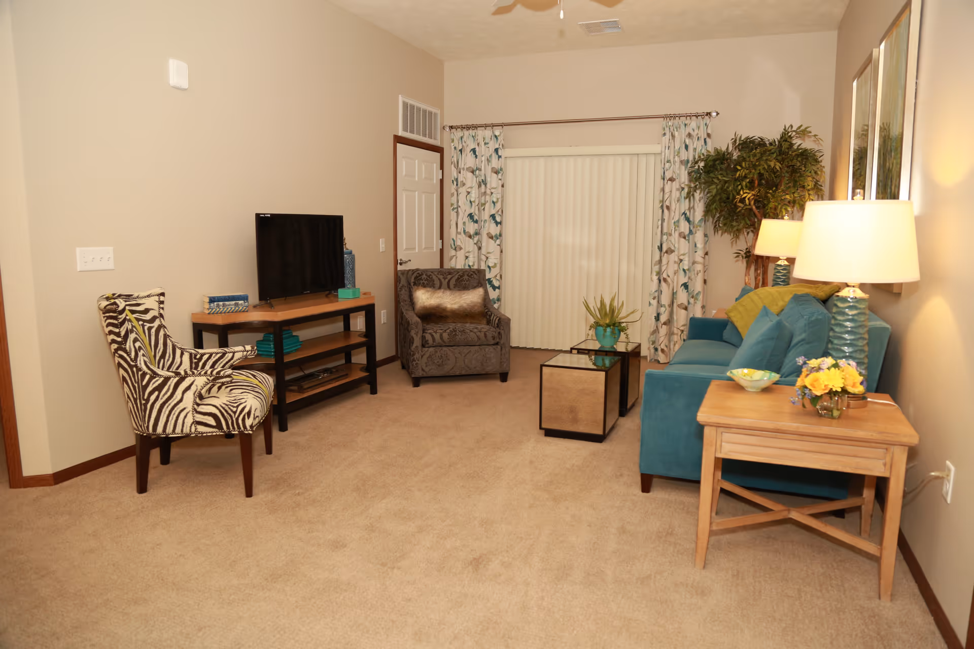A cozy living room with beige carpet and light beige walls. The room features a teal sofa with green and teal cushions, a wooden side table with a lamp and a small flower arrangement, a patterned armchair, a zebra-print chair, a TV on a wooden stand, a small glass coffee table with a plant, and floral curtains covering a sliding glass door. There is also a large potted plant in the corner and a ceiling fan above.
