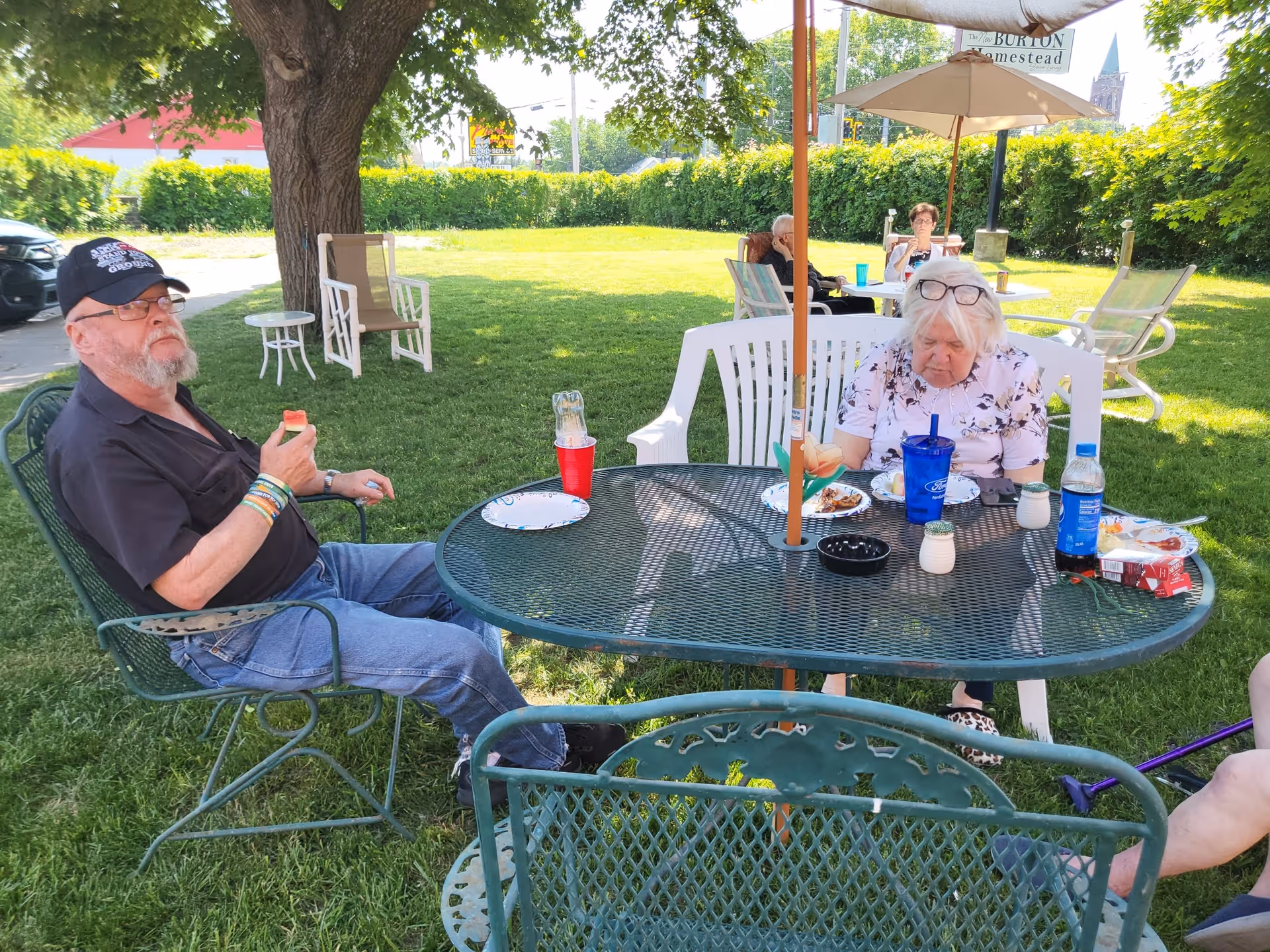 Several elderly people sitting outdoors at metal and plastic tables with umbrellas, enjoying food and drinks on a sunny day with green grass and trees around.