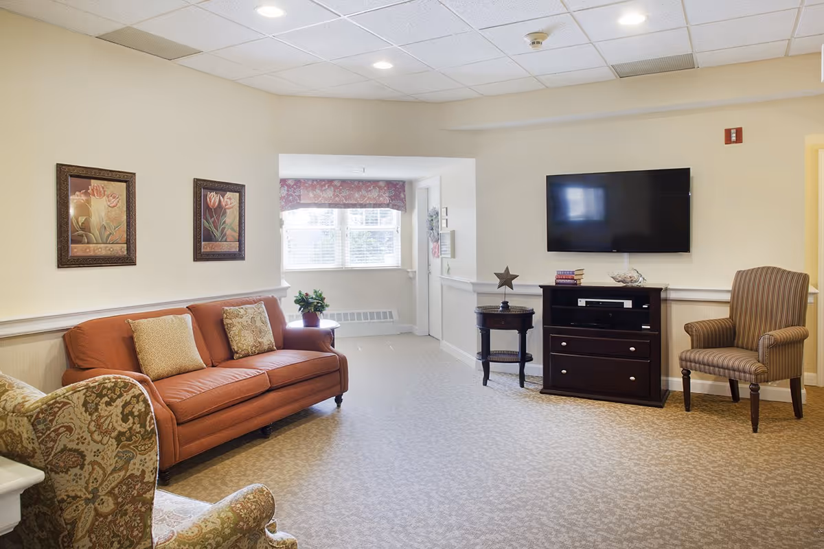 A bright communal living room with an orange sofa, patterned armchairs, a wall-mounted TV above a dark wood console, framed floral prints, and a window.