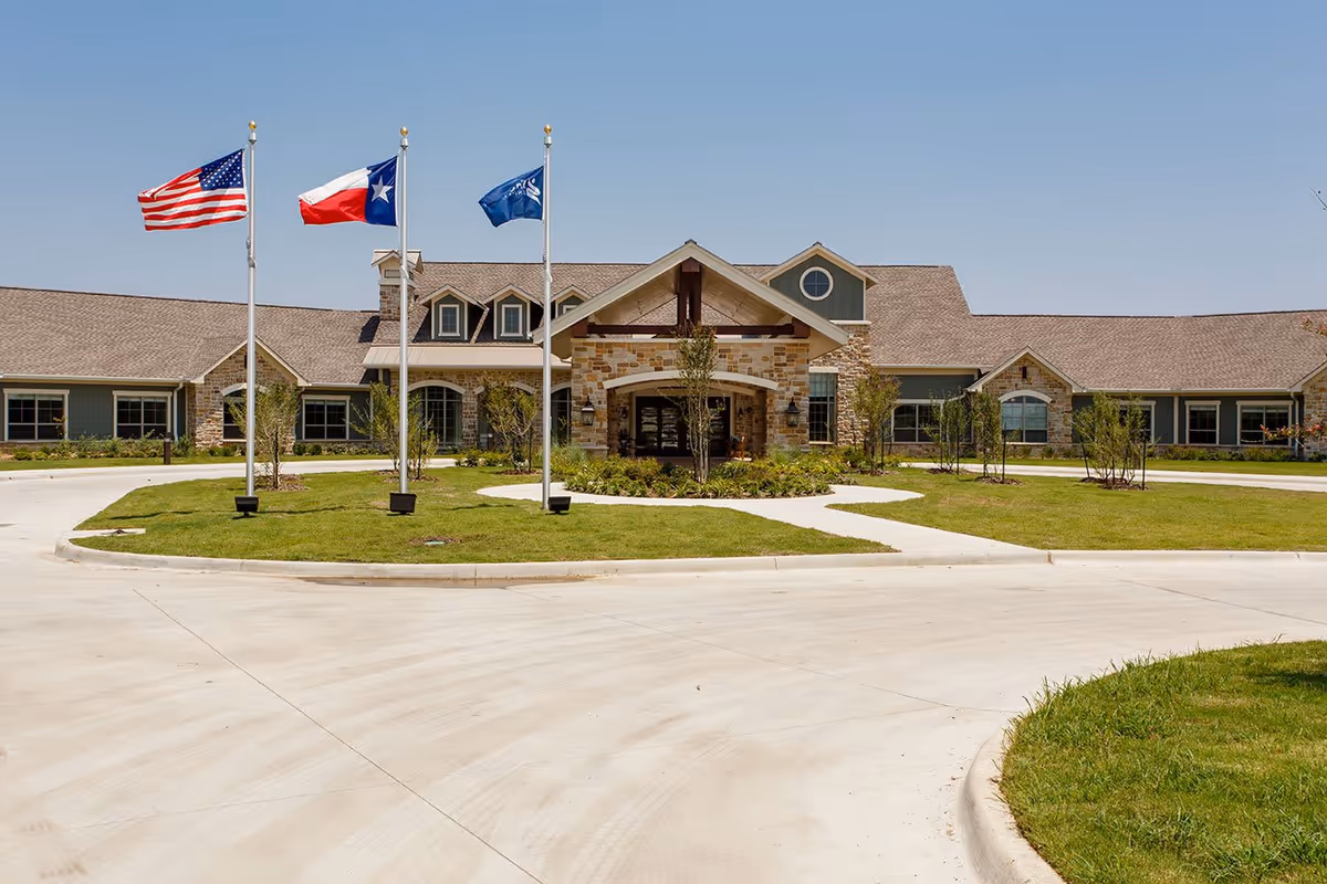 Front exterior view of Heritage Place Assisted Living & Memory Care building with a circular driveway, three flagpoles displaying the American flag, Texas state flag, and another flag, and a clear blue sky.