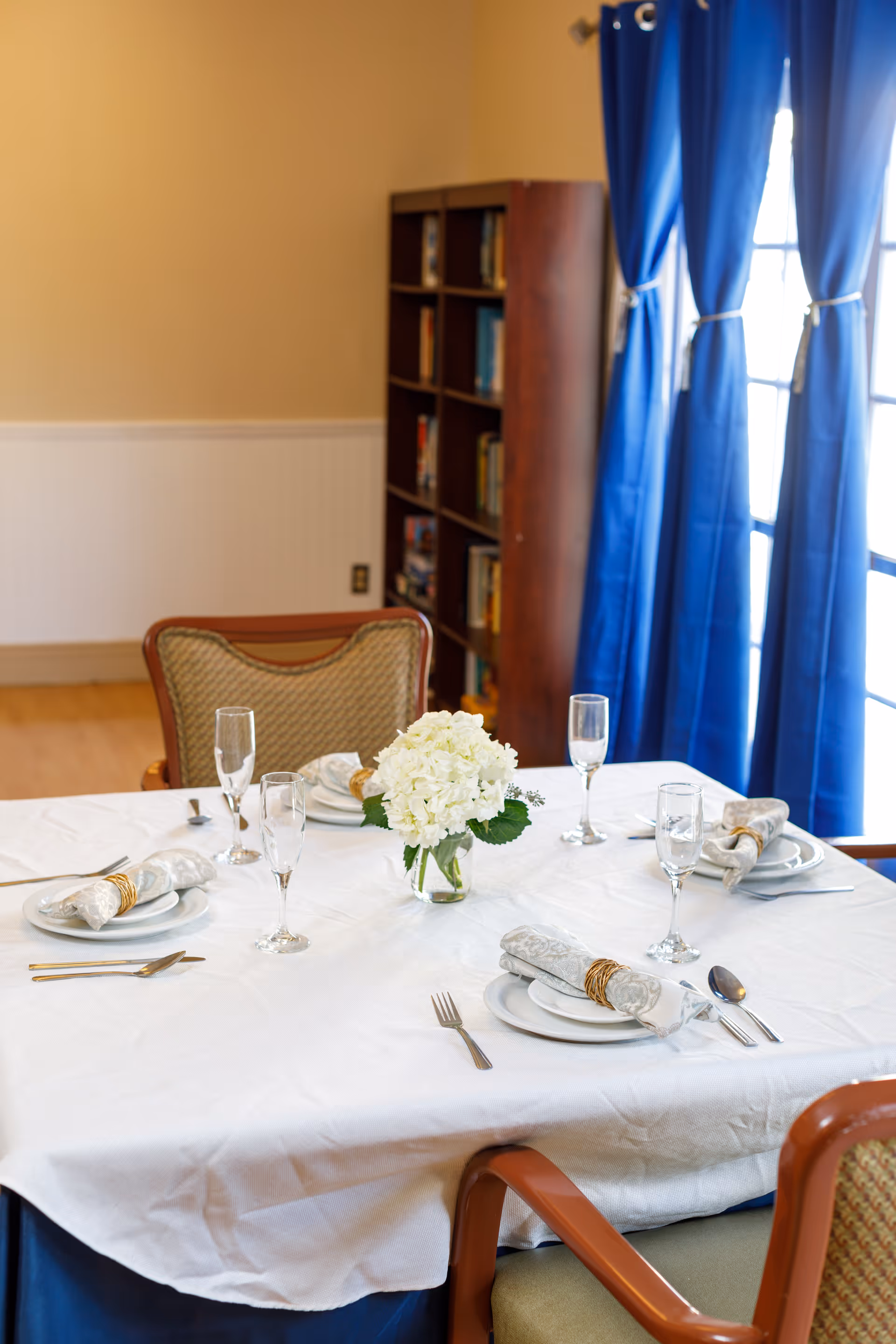 A dining table set for four with white tablecloth, napkins with rings, plates, cutlery, and empty champagne glasses. A small vase with white flowers is in the center. In the background, there is a wooden bookshelf filled with books and blue curtains covering windows.