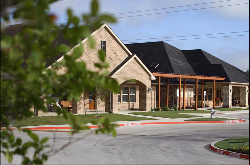 Front exterior of a single-story brick senior living building with a covered entrance and driveway, partially framed by leafy branches.