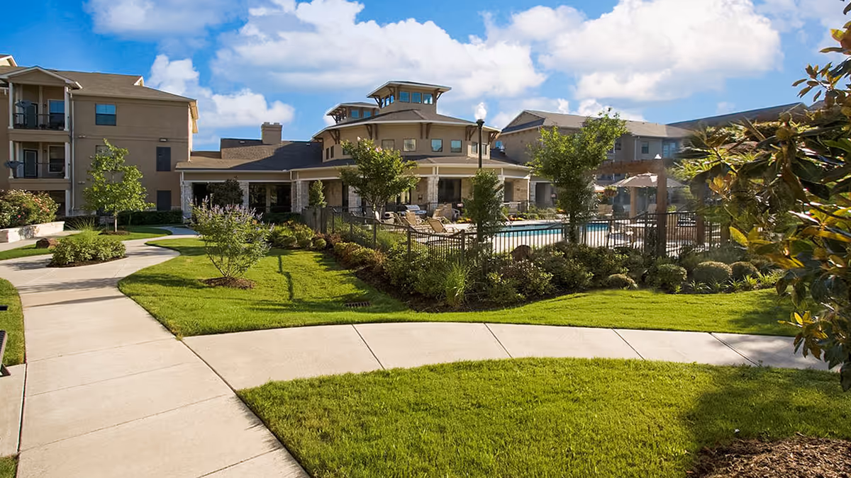Landscaped courtyard with curved walkways, green lawns, a fenced pool and surrounding apartment-style buildings under a blue sky.