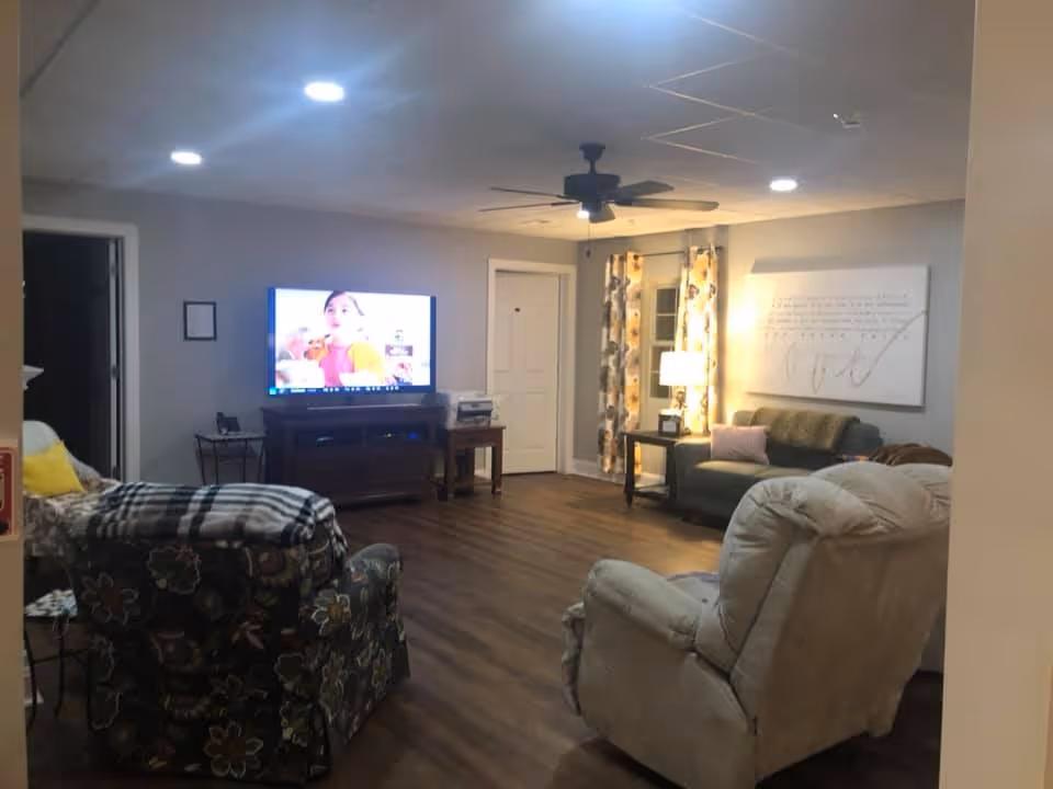 A cozy living room with wooden flooring, a ceiling fan, and recessed lighting. The room features a large flat-screen TV on a wooden stand, a floral patterned armchair with a blanket draped over it, a beige recliner, and a green sofa with a pink pillow. There is a side table with a lamp next to the sofa, and floral curtains hang by a door in the corner.