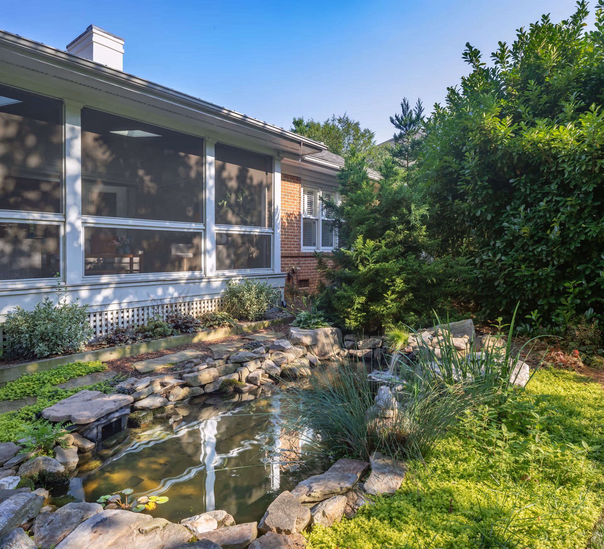 A screened porch and brick wall beside a landscaped garden pond surrounded by rocks and greenery.
