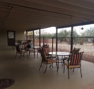 Covered outdoor patio area with several round tables and metal chairs with cushions, overlooking a fenced yard with desert landscaping and trees.